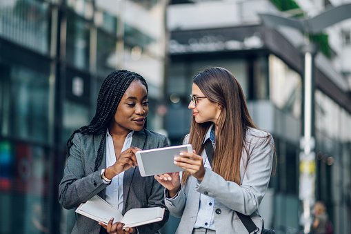 Two diverse female coworkers standing outside in front of office buildings discussing business plan and looking at a digital tablet. Leadership concept. Copy space.