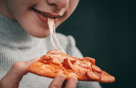 Happy caucasian girl holding a slice of pizza with stretching mozzarella cheese in her hand and biting her mouth on a black background in focus, close-up side view.