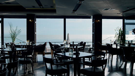 Served lunch tables in spacious stylish cafeteria with beautiful sea view. Comfortable hotel restaurant dining hall background. Modern panoramic sky bar dark interior. Luxury dinner concept.