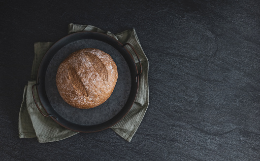 One round rye bread in a vintage metal plate with a kitchen napkin lies on the left on a black stone background with copy space on the right, flat lay close-up. Bread baking concept.