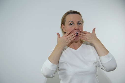 adult woman 40-50 years old covers her mouth with her hands Do not say She is surprised mouth open eyes wide open raised eyebrows blond hair indignant white clothes