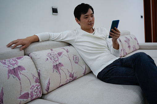 The man is sitting on the sofa drinking water and using his mobile phone