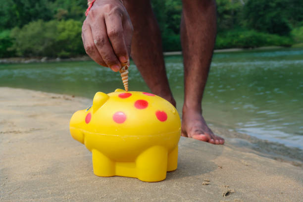 Stock photo showing close-up view of plastic, yellow piggy bank with red spots on sandy beach, at sea water's edge at low tide as an unrecognisable person places a spiral seashell into the slot on top of the piggy bank. Holiday finances and savings concept.