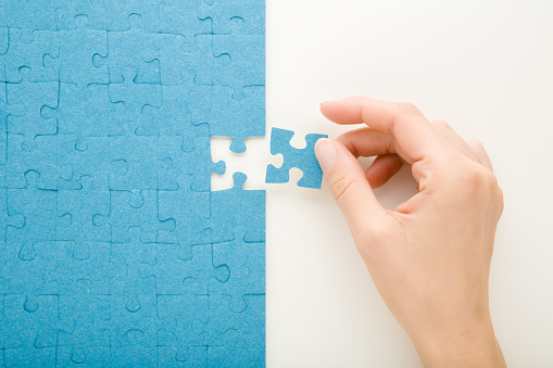 Young adult woman hand holding and putting last one blue puzzle piece on white table background. Closeup. Top down view.