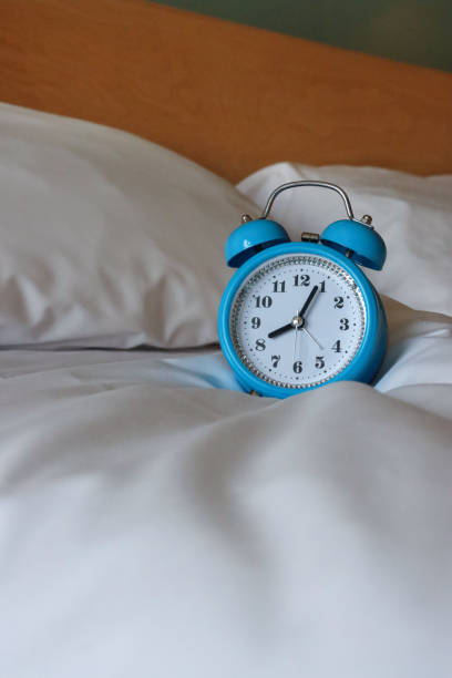 istock Image of blue metal case, retro alarm clock, double bell, analogue display clock with white dial sitting on white duvet bed cover, black numerals, hour and minute hands, pillows leaning on wood and glass headboard, focus on foreground, copy space 1456312663