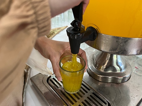 A woman hand wearing brown cloth pouring orange juice from dispenser into a small glass.