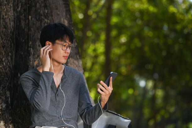 istock Young asian man using smart phone while sitting under tree shade on sunny day surrounded by nature. 1456317869