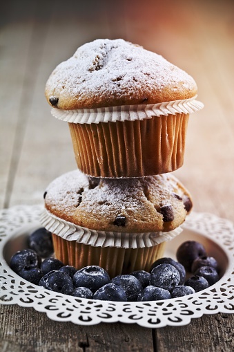 Two homemade fresh muffins with sugar powder and blueberries on white plate on rustic wooden table background.