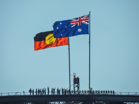 The Australian and Australian Aboriginal flags fly on the Sydney Harbour Bridge on a windy and sunny afternoon in summer, with silhouettes of people climbing the bridge. Some have raised their hands, perhaps for a photo taken by the guide.  This image was taken from Mrs Macquarie's Chair on 14 January 2023.