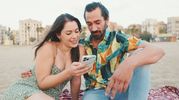 istock Young couple uses mobile phone while sitting on the beach on buildings background, close-up 1456353039