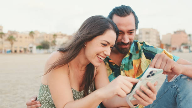 istock Young couple uses mobile phone while sitting on the beach on buildings background, close-up 1456353055
