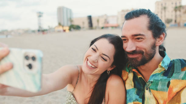 istock Happy couple taking selfie on mobile phone while sitting on the beach 1456353088