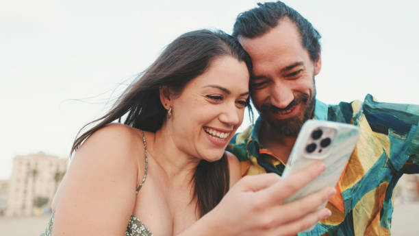 istock Young couple uses mobile phone while sitting on the beach on buildings background, close-up 1456353090