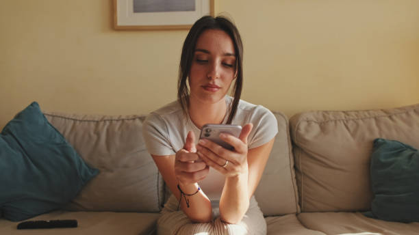 istock Young woman using mobile phone for texting and scrolling while sitting on sofa at home 1456355347