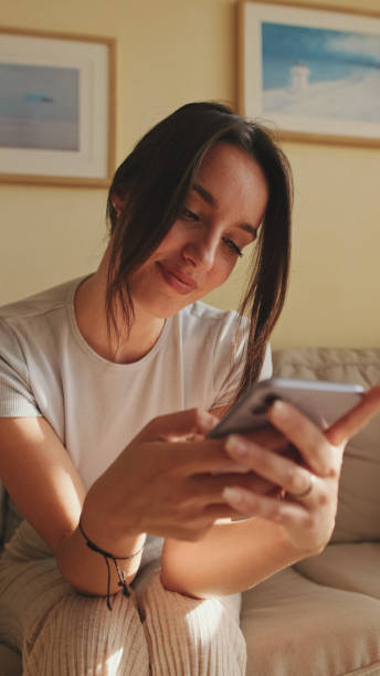 istock Young woman using mobile phone for texting and scrolling while sitting on sofa at home 1456355367