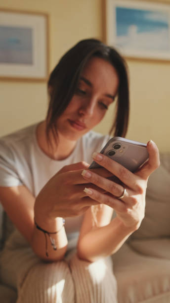 istock Young woman using mobile phone for texting and scrolling while sitting on sofa at home 1456355375