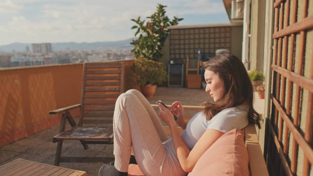 istock Young woman using mobile phone while sitting on balcony at home 1456355436