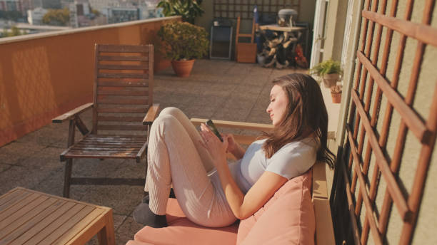 istock Young woman using mobile phone while sitting on balcony at home 1456355481