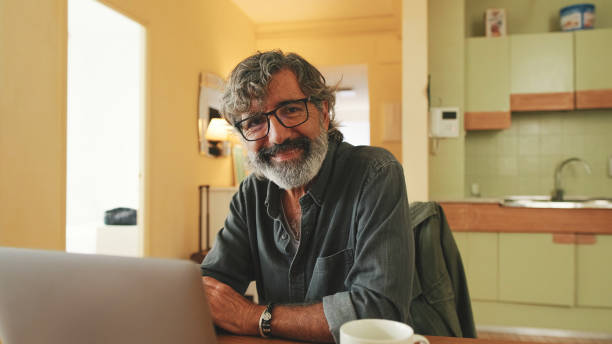 istock Senior working on laptop sitting in home kitchen and loooks at on camera 1456355962