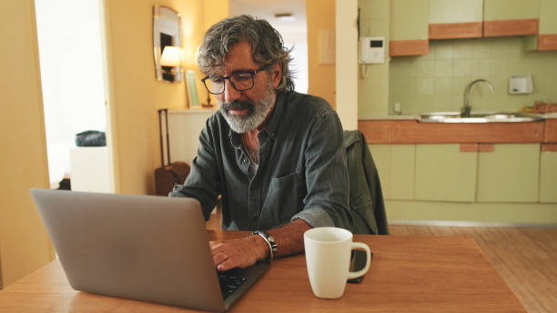 istock Elderly positive man typing on laptop sitting in home kitchen 1456356001