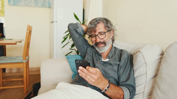 istock Middle-aged man relaxing on his couch while in home and using his smartphone to text 1456356034