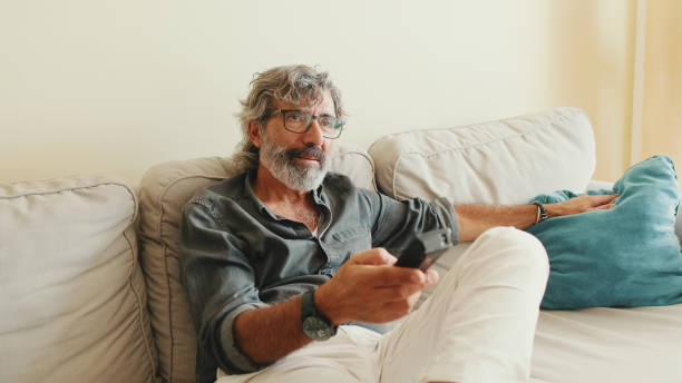 istock Close-up of the hand of an elderly man with remote control, changes channels while sitting on the couch at home 1456356065