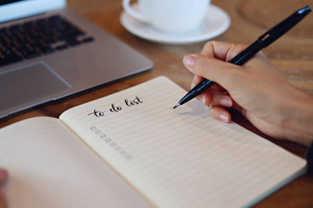 ペンを持つ手、オフィスの机の上のノート、ラップトップ、コーヒーカップにリストを計画 - やることリスト ストックフォトと画像