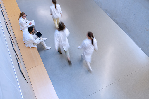 Multi-racial group of chemistry students in white uniforms during lectures in university corridors while casually chatting. View from above. Blurred figures in motion.
