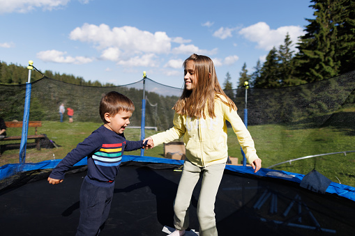 An older sister with flowing long hair in a yellow tracksuit and her little brother in a cute blue suit are jumping together on a netted trampoline in the backyard in the spring at the weekend