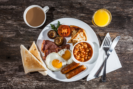 Full fried English breakfast with fried eggs, sausage, bacon, beans, potato bread, mushrooms and tomato on a white plate. Served with orange juice and coffee.