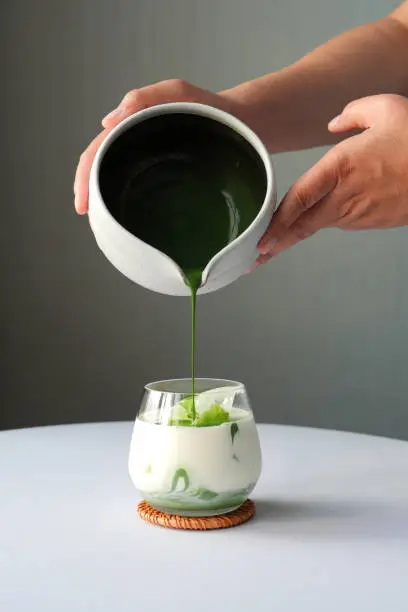 Photo of Preparing matcha latte green tea with milk in a glass on white table over gray background, hand hold a bowl pouring matcha to the glass with ice cubes.