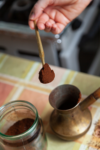 We brew natural coffee. Pour ground coffee into a brass turk with a spoon on a female hand. There is a glass jar nearby