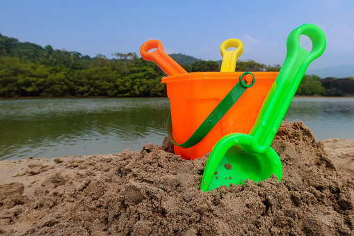 Stock photo showing a collection of multicoloured, plastic beach toys on sandy beach at water's edge of sea. Toys include a bucket, spades, a rake and a watering can in front of backdrop of woodland with palm trees.