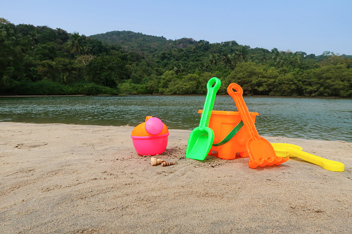 Stock photo showing a collection of multicoloured, plastic beach toys on sandy beach at water's edge of sea. Toys include a bucket, spades, a rake and a watering can in front of backdrop of woodland with palm trees.