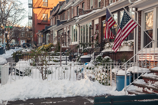 The photo shows a snow-covered residential area of Brooklyn. Row houses entrances. Front door lantern, Christmas decoration and American flag are on the foreground. Winter storm ended early morning today, snow formed drifts. Cars parked at the sides of the street are also covered with snow. Midday. December 17, 2020. NYC. Brooklyn. Bay Ridge. New York. USA