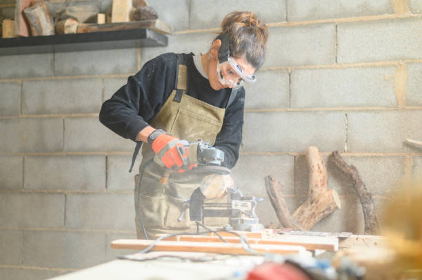 giovane donna falegname che lucida il legno con la macchina sul posto di lavoro. giovane donna falegname in occhiali che lavora con lucidatore sul posto di lavoro. - drill power tool work tool carpenter foto e immagini stock