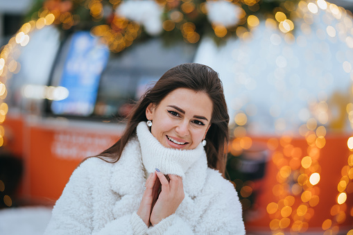 Adorable European brunette girl in white sweater and white fur coat toothy smiles looks at camera stands at outdoor food court against blurred garlands. Girl on winter holidays. Christmas, new year.
