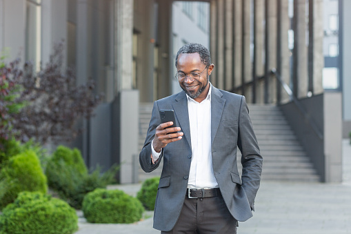 Successful african american businessman outside modern office building in business suit using smartphone, mature boss checking message smiling and reading news online from phone.