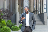 Successful african american businessman outside modern office building in business suit using smartphone, mature boss checking message smiling and reading news online from phone