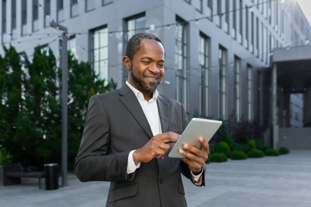 istock Successful african american man in business suit smiling and using tablet computer, businessman outside modern office building walking 1456419394