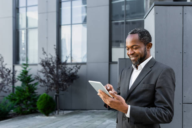 istock Successful african american man in business suit smiling and using tablet computer, businessman outside modern office building walking 1456419400