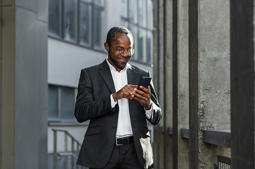 Happy successful African American businessman boss walking outside office building, manager holding smartphone reading message and smiling, using communication app.