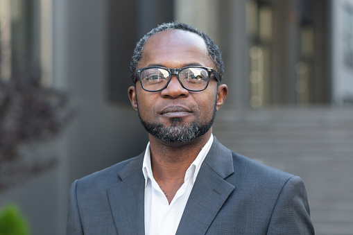 Close up portrait of serious businessman, afro american man in business suit and glasses looking at camera thinking, boss outside office building.