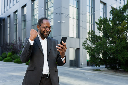 African american businessman boss outside modern office building using phone, senior man celebrating victory success reading good news online from smartphone, holding hand up gesture of triumph .