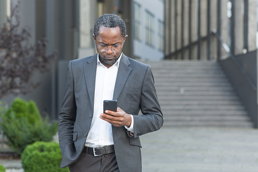Serious and successful African American boss businessman outside office building using smartphone, man in business suit reading news online thinking and browsing internet pages.