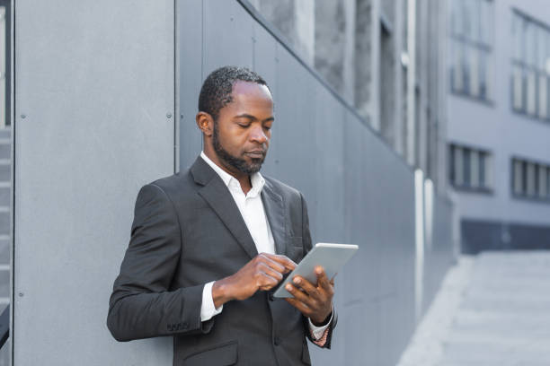 istock Serious thinking african american businessman using tablet computer, man in business suit mature reading news online standing outside office building 1456419552