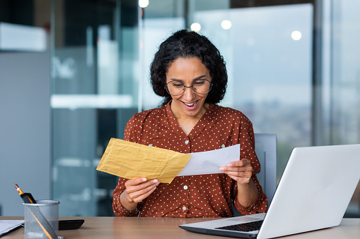 Happy young Latin American woman received bill with earned salary, cash reward, good news. He is sitting at the desk in the office, holding an envelope with a letter in his hands.