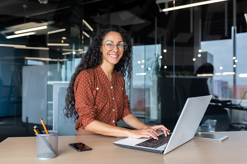 Young beautiful hispanic woman working inside modern office, businesswoman smiling and looking at camera at work using laptop.