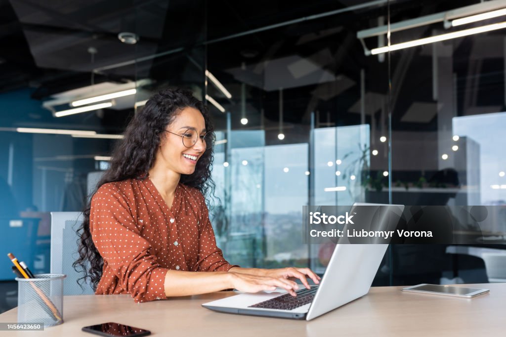 Happy and smiling hispanic businesswoman typing on laptop, office worker with curly hair and glasses happy with achievement results, at work inside office building Happy and smiling hispanic businesswoman typing on laptop, office worker with curly hair and glasses happy with achievement results, at work inside office building. People Stock Photo