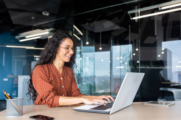 Happy and smiling hispanic businesswoman typing on laptop, office worker with curly hair and glasses happy with achievement results, at work inside office building Happy and smiling hispanic businesswoman typing on laptop, office worker with curly hair and glasses happy with achievement results, at work inside office building. using-laptop stock pictures, royalty-free photos & images